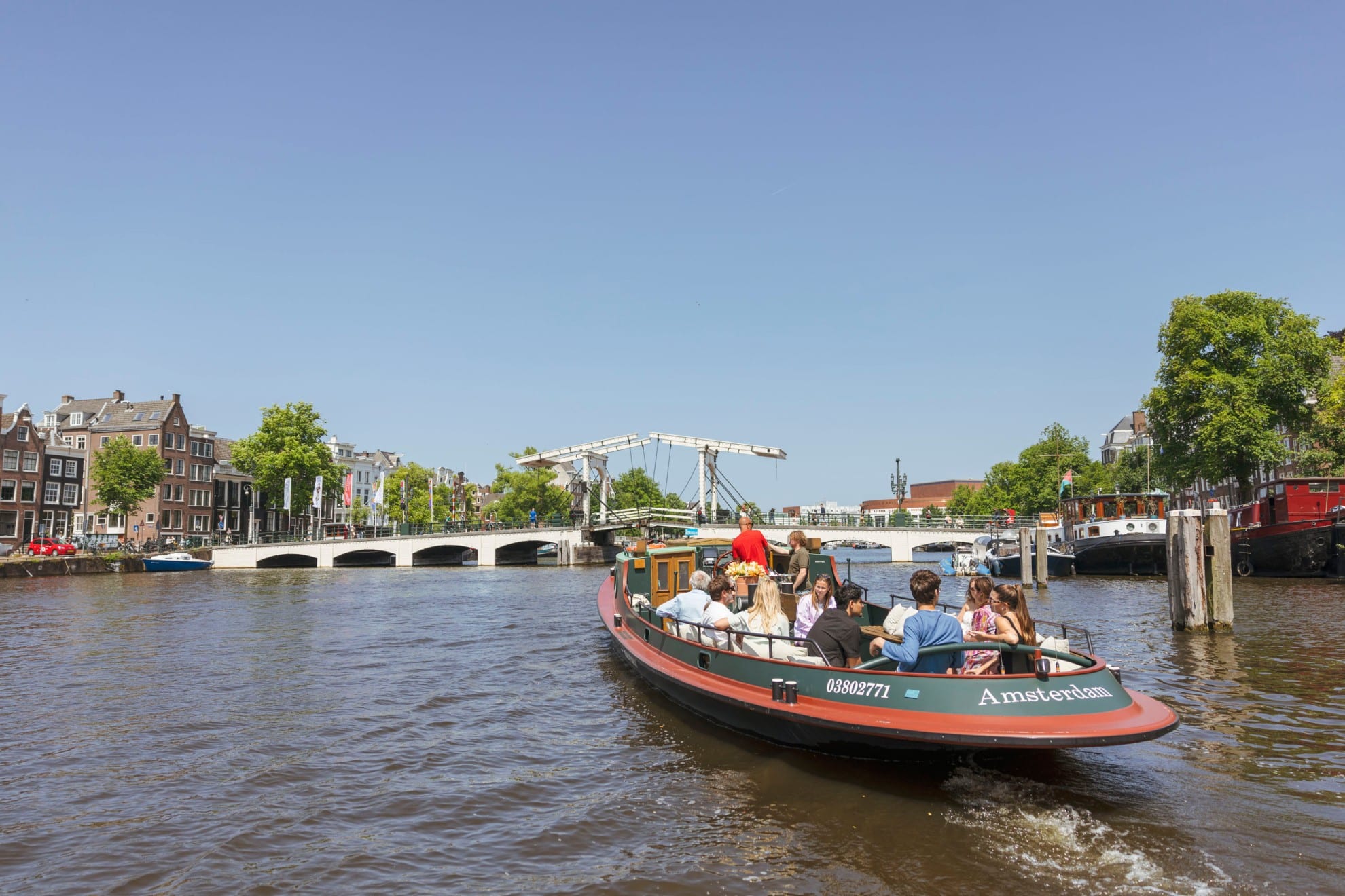 Een klassieke rondvaartboot vol passagiers vaart over de Amsterdamse gracht richting de Magere Brug op een zonnige dag.