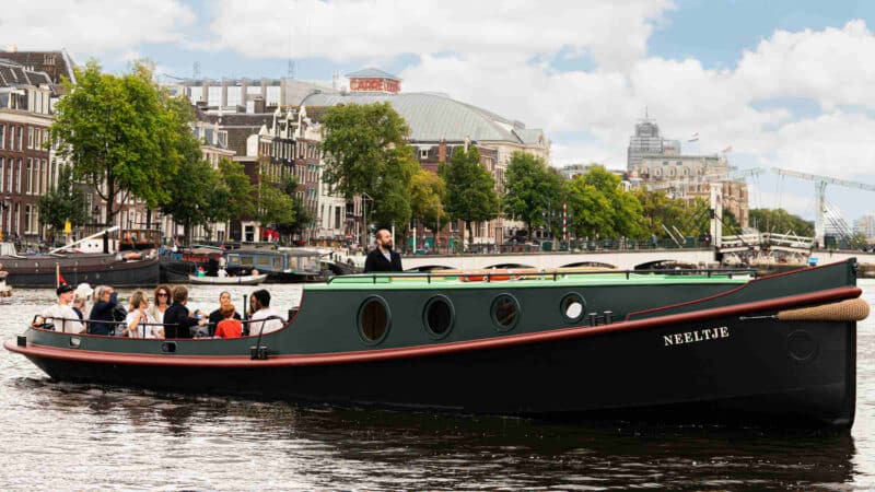 Een rondvaartboot met passagiers vaart onder een stenen boogbrug door op de Amsterdamse grachten, terwijl voetgangers over de brug lopen.