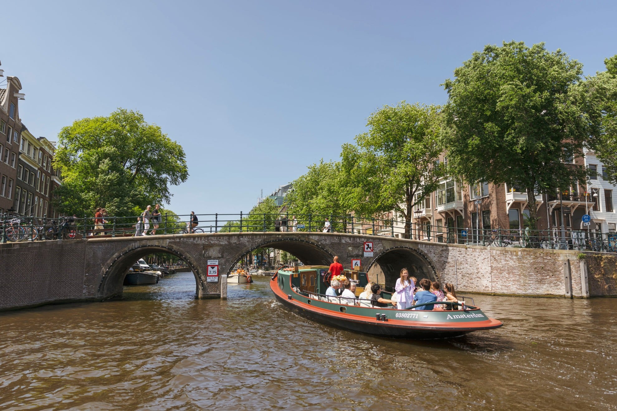 Bootjehureninamsterdam.com – Rondvaart op een klassieke sleepboot Ein Ausflugsboot mit Passagieren fährt unter einer Steinbogenbrücke über die Amsterdamer Grachten hindurch, während Fußgänger über die Brücke gehen.